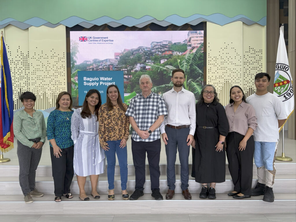 Diverse GCIEP team stands in a conference venue in front of a digital display showing the Baguio Water Supply Project.